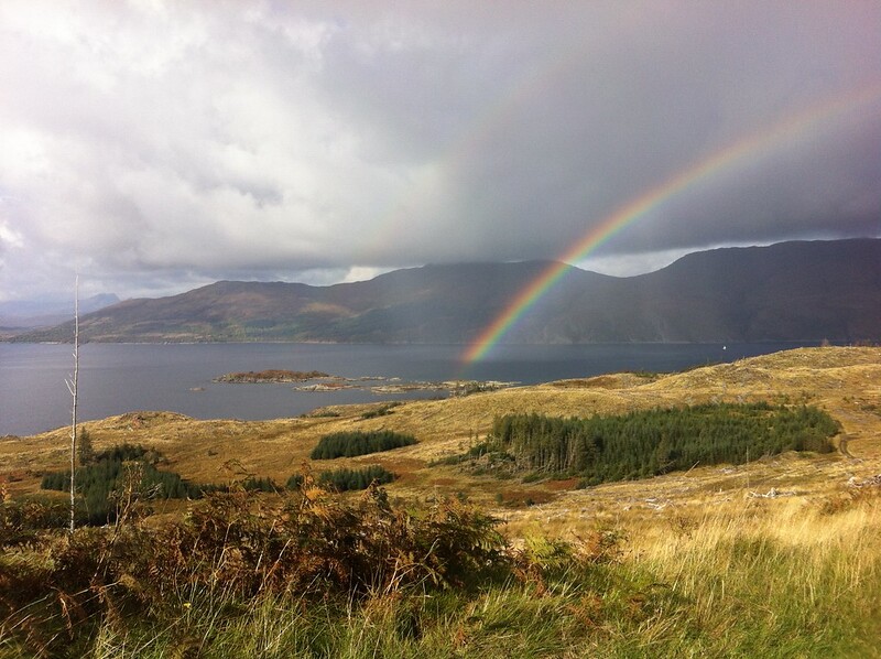 Grassy hill sloping down to sea with mountains to rear and rainbow