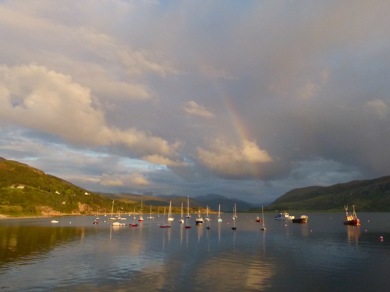 Small boats in harbour with rainbow above sea and hills behind 