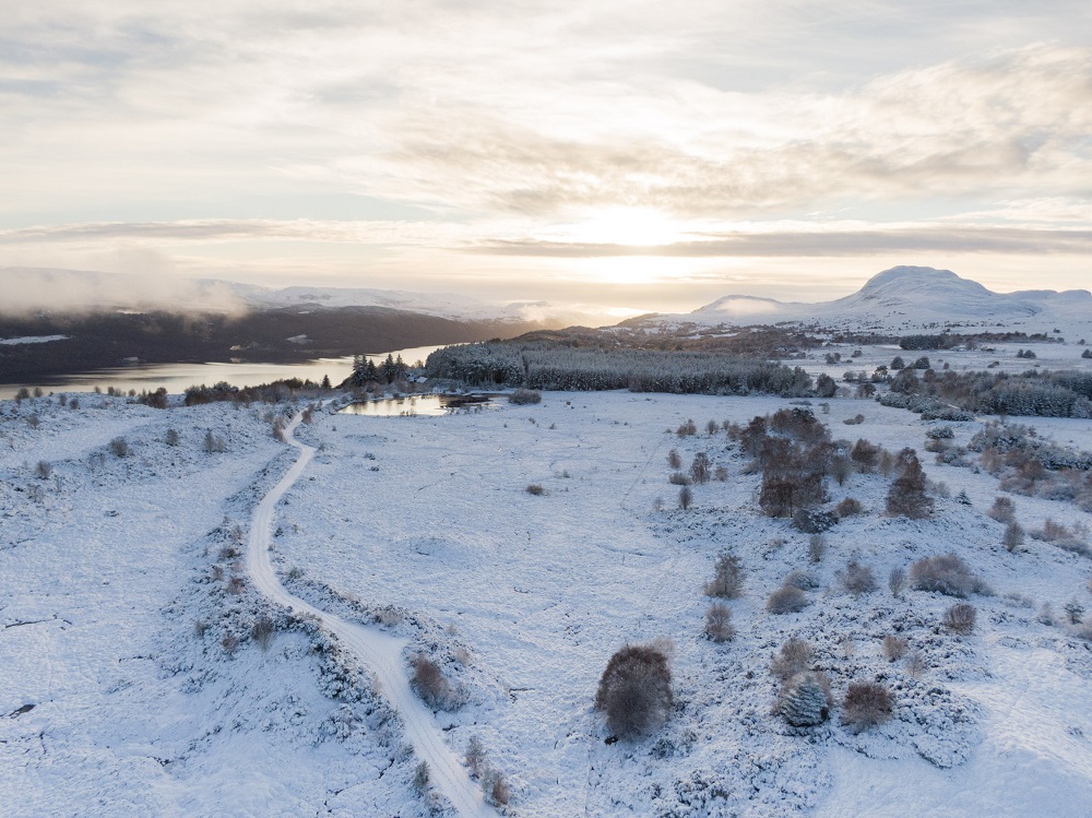 Road running through snow-covered landscape of fields, lochs and mountains to rear 