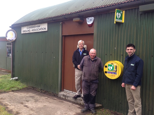 Picture of a WAT IF? trustee and a Muirhall Energy representative outside an Angling club which has a defibrillator on wall