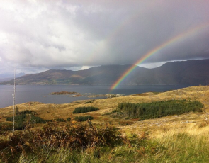 Grassy hill sloping down to sea with mountains to rear and rainbow