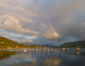 Small boats in harbour with rainbow above sea and hills behind 
