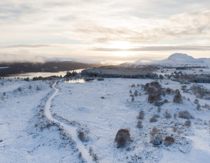 Road running through snow-covered landscape of fields, lochs and mountains to rear 