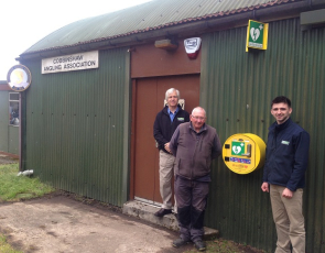 Picture of a WAT IF? trustee and a Muirhall Energy representative outside an Angling club which has a defibrillator on wall
