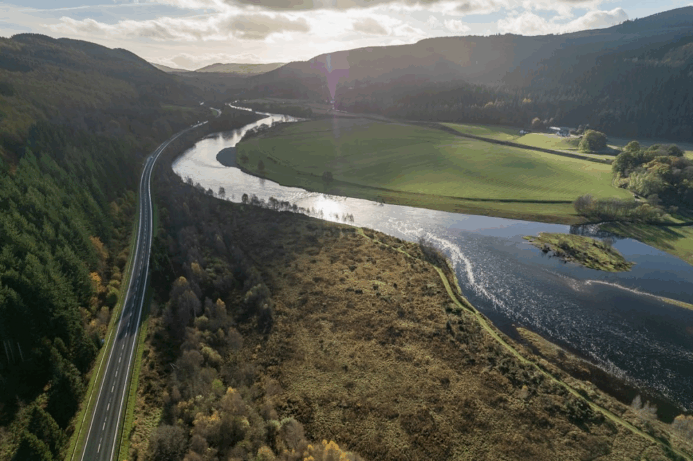 A winding river flows through a lush valley with green fields and dense forests under a partly cloudy sky.