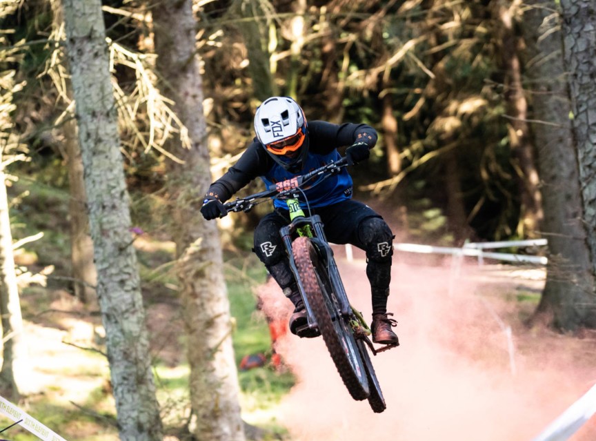 Mountain biker wearing a white helmet and orange goggles jumps mid-air on a forest trail surrounded by trees and dust.