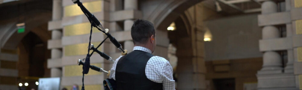 Man in a checked shirt and black waitscoat playing bagpipes inside a historic stone building with arches.