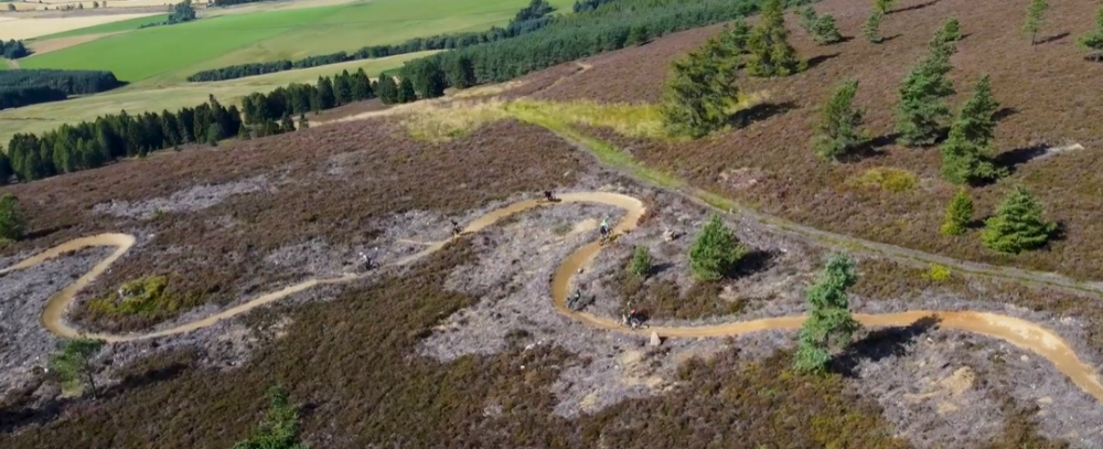 ariel shot of green agricultural land in the distance becoming more brown with sparse vegetation in the foreground. Mountain bike track with cyclist is also in the foreground