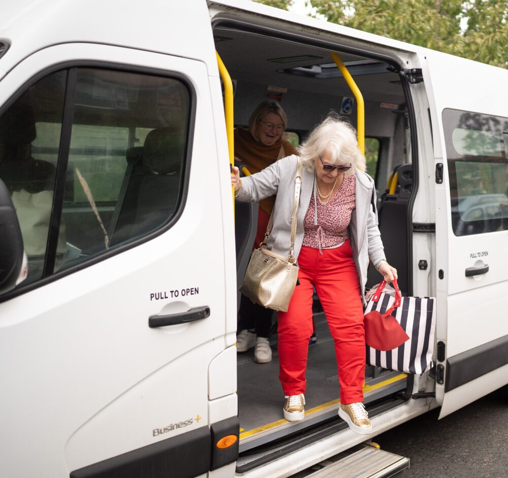 woman in red trousers and top stepping down form a white mini bus