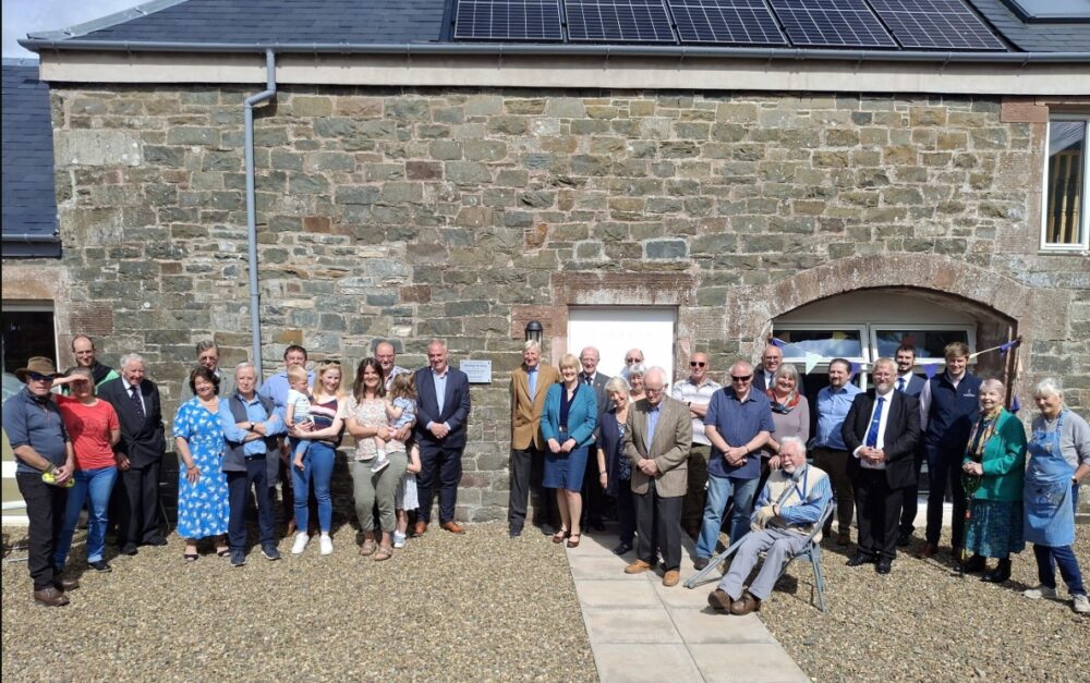 Large group of people gathered outside a stone building with solar panels on the roof on a sunny day.