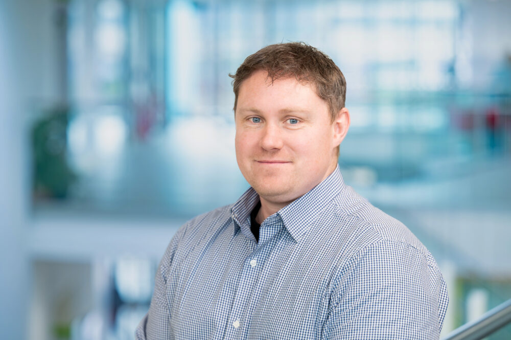 Man wearing a blue and white checkered button-up shirt standing indoors with a blurred modern office background.