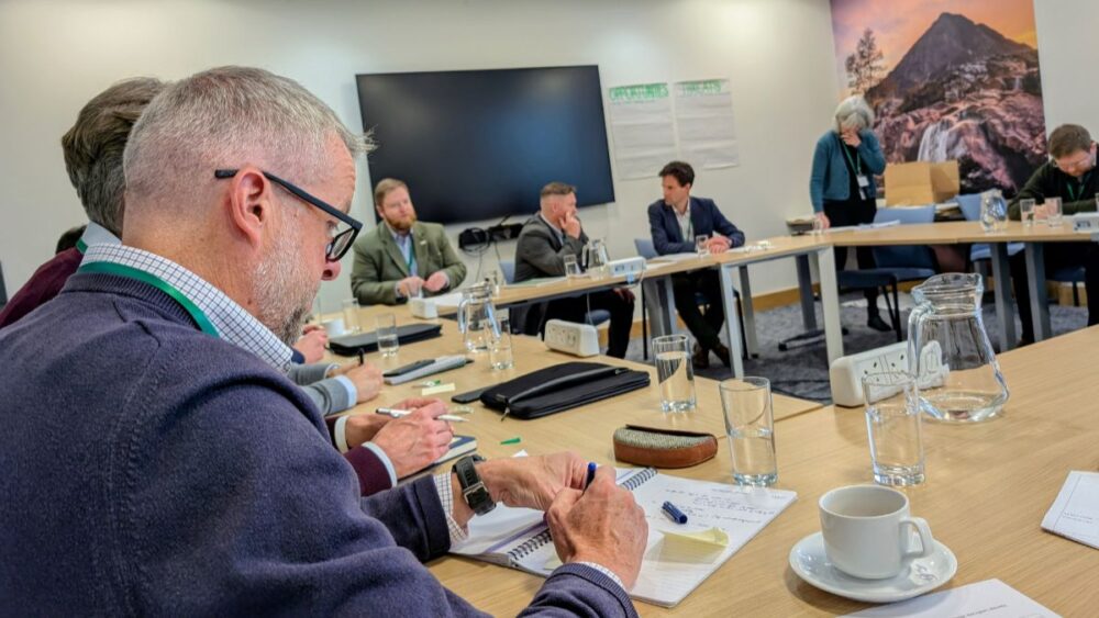 Group of professionals seated around a U-shaped conference table during a meeting with notebooks, glasses of water, and a large screen on the wall.