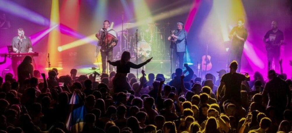 Live band performing on stage with colorful spotlights illuminating the musicians and a large crowd enjoying the concert.