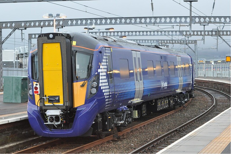Modern blue and yellow passenger train stopped at a curved platform under overhead electric wires on a cloudy day.