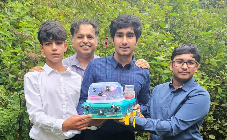 Four people in casual shirts holding a technology project robot with various sensors in a leafy environment.