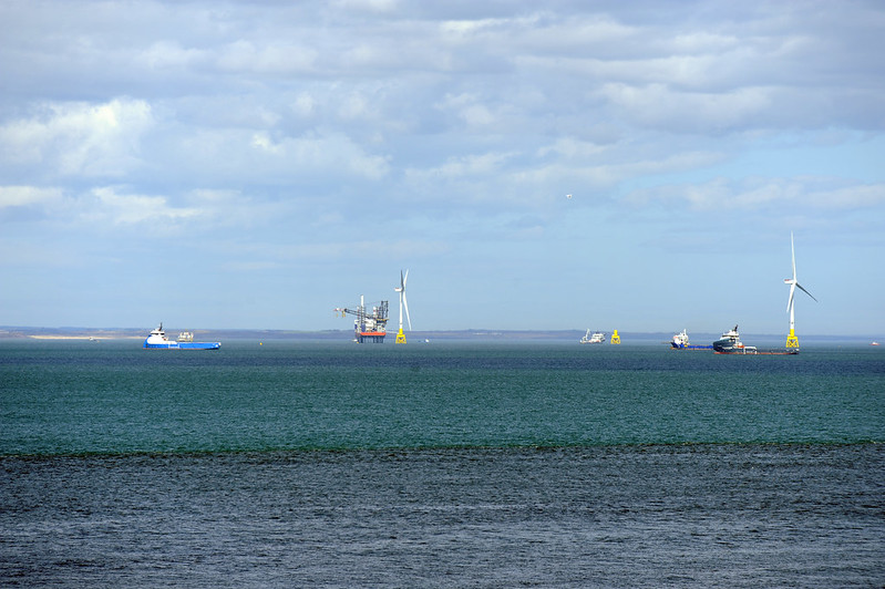 Offshore wind turbines and service vessels operating near an oil platform under a cloudy sky.