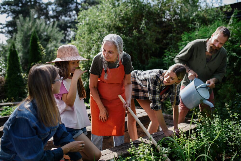Group of people gardening together, one wearing an orange apron using a hoe, surrounded by lush green plants in a sunny garden.