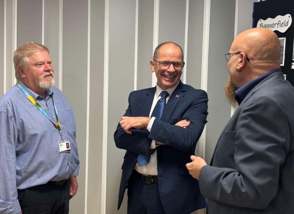 Three men talking in front of a noticeboard in a room with striped walls.