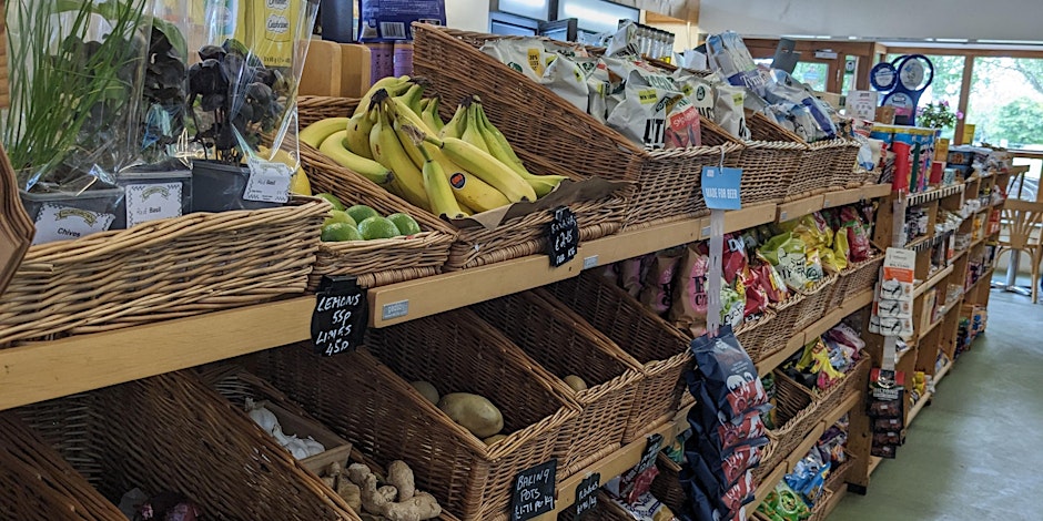 Wooden shelves in a grocery shop display fresh bananas, limes, herbs, and various snack bags in wicker baskets under natural light.