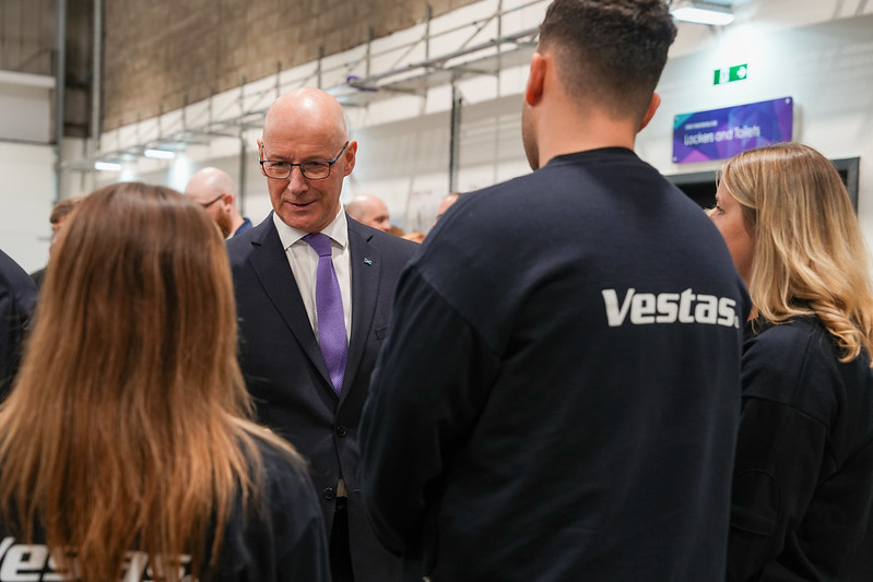 A man in a suit and purple tie speaks with three people wearing black Vestas shirts inside an industrial facility.