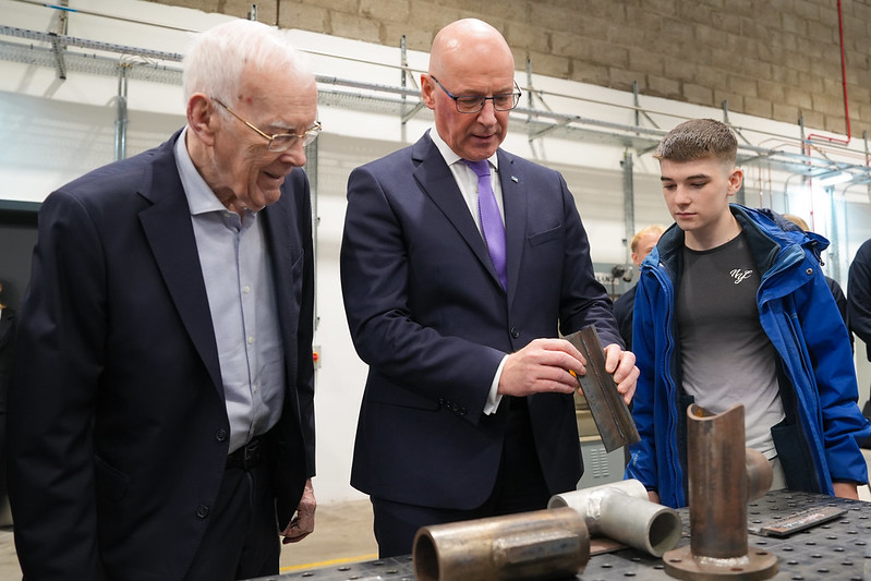 Three men in a workshop examining metal pipe components on a workbench, with industrial equipment in the background.