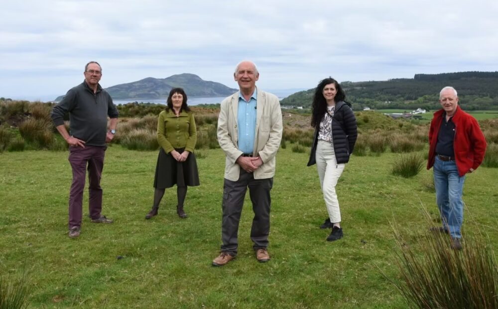 Five people standing spaced apart on a grassy field with hills and water in the background under a cloudy sky.