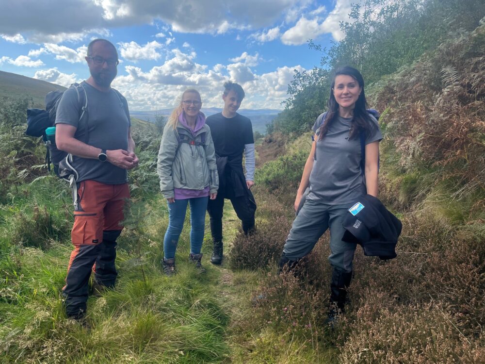 Group of four hikers standing on a grassy trail surrounded by hills and cloudy blue sky.