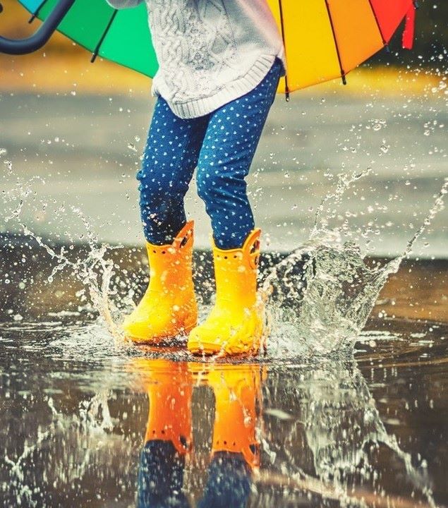 Child with yellow wellies and a multicoloured umbrella jumping in a puddle