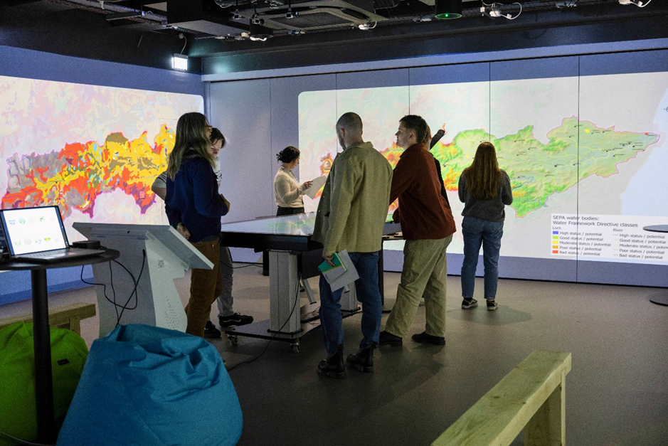 Group of people interacting with large digital maps projected on walls in a modern, dimly lit room with touchscreen tables.