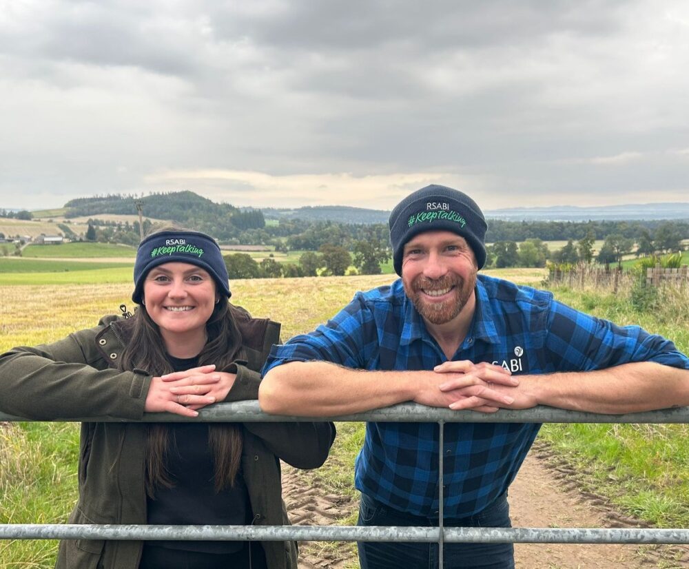 Two people wearing RSABI beanies lean on a metal gate with rolling green fields and hills in the background under a cloudy sky.