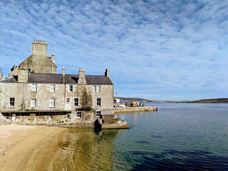 Old multi-story stone house beside clear water with a sandy shore and distant harbour structures.