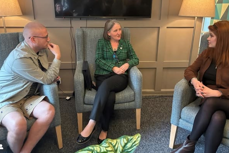 Three people seated in a cosy room with grey armchairs, engaged in conversation under warm lighting and a wall-mounted TV.