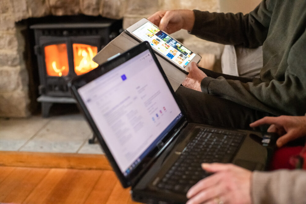 Two people sitting near a cosy fireplace using a laptop and a tablet for browsing and research indoors.