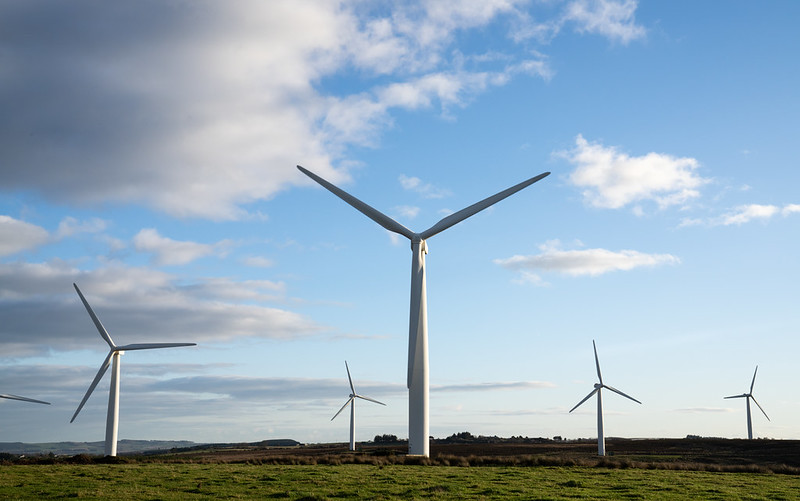 Several large white wind turbines stand on a grassy field under a partly cloudy blue sky during daylight.