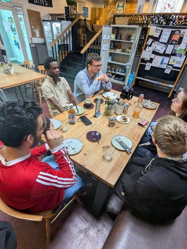 Group of five people sitting around a wooden table in a cozy cafe with plates, drinks, and condiments scattered on the table.