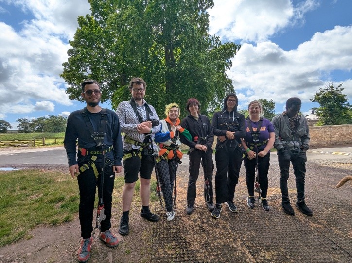 Group of seven people standing outdoors in safety harnesses and gear, ready for an adventure activity under a partly cloudy sky.