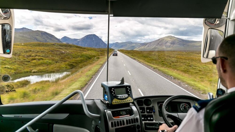 Bus driver wearing sunglasses steering on a rural road surrounded by grassy fields and mountainous landscape in the background.