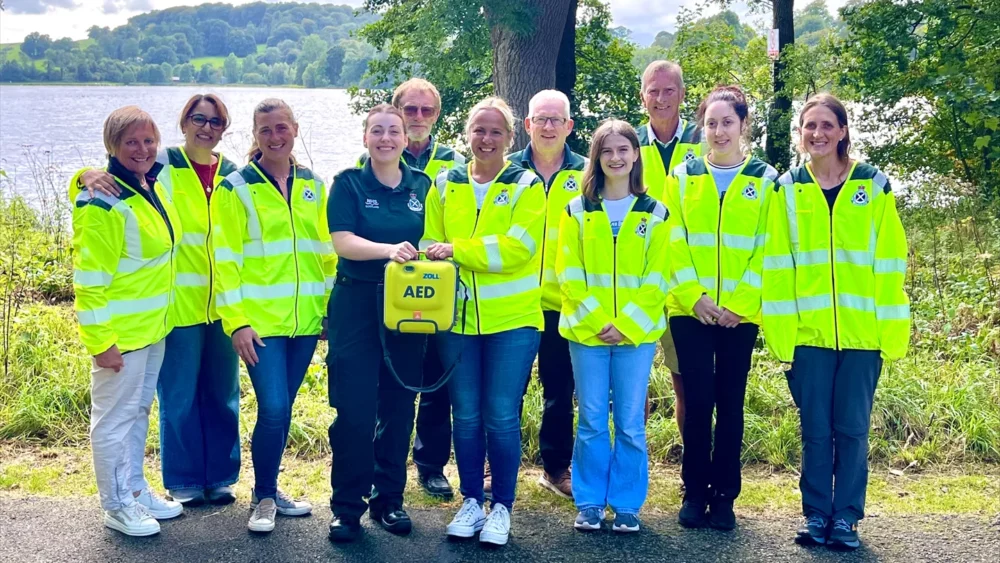 Group of volunteers wearing bright yellow safety jackets standing outdoors near a lake with trees in the background.