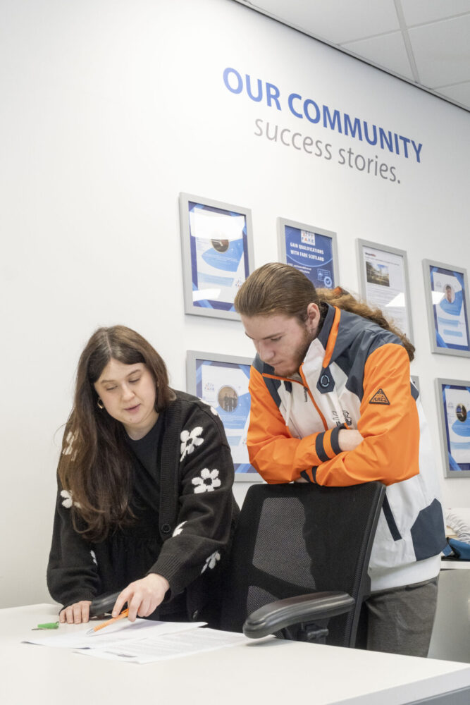 A woman and a man review paperwork together in a bright room decorated with framed community achievement posters.