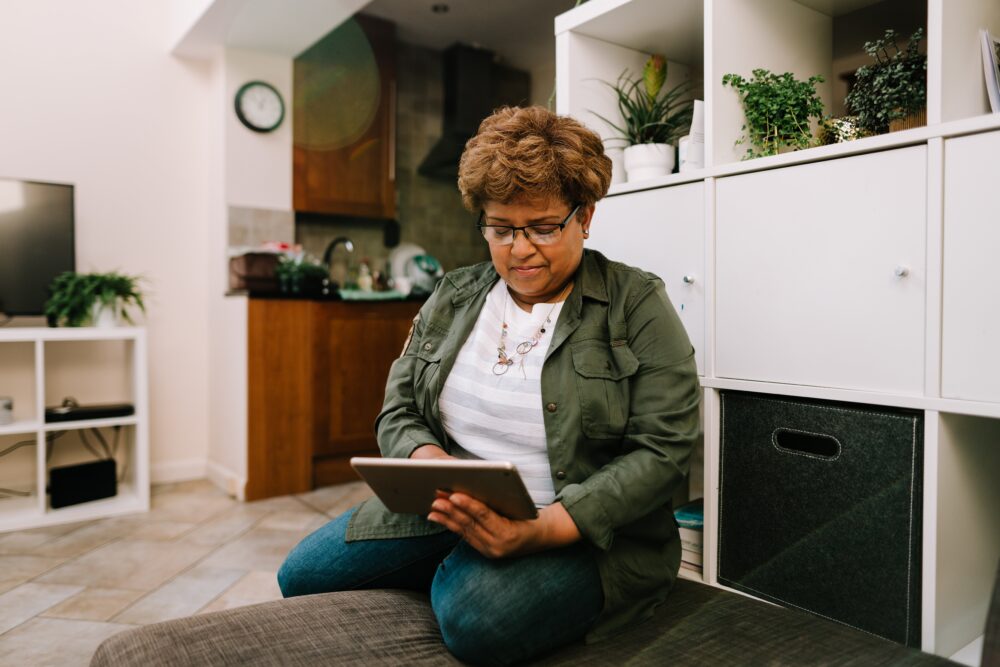 Person sitting on a couch in a cosy living room, using a tablet with a kitchen visible in the background.