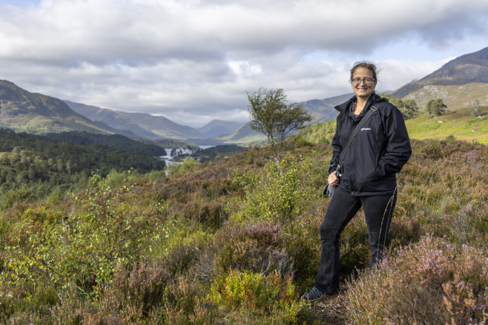 Woman standing in heather on a hillside with mountains in background