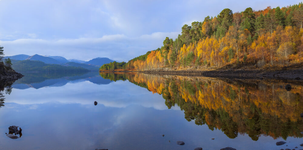 Serene water mirrors colourful autumn foliage on a forested hillside with distant mountains in the background.