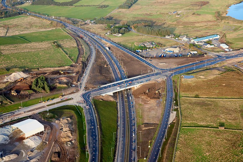 Overhead shot of a complex road junction featuring several lanes, vehicles, and adjacent open fields under clear weather.