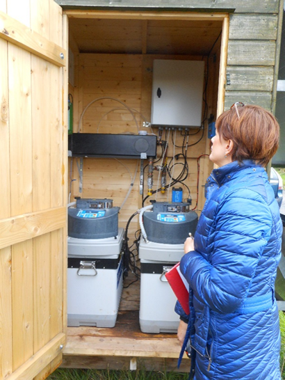 Person in blue jacket inspecting water monitoring units inside a wooden shed with open door.