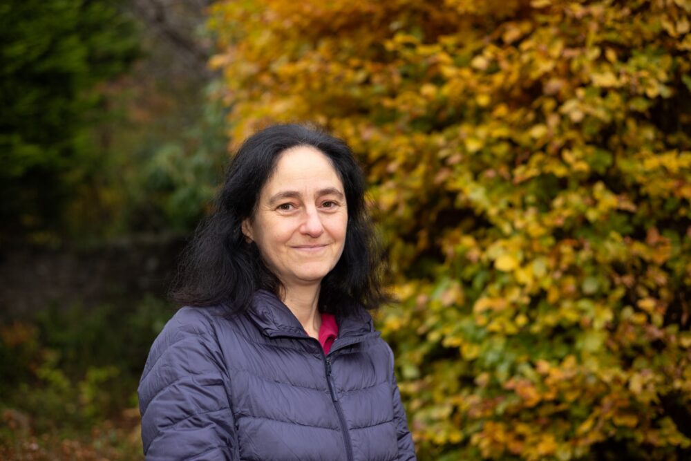 Women with black hair and wearing a blue jacket standing in front of trees in autumn