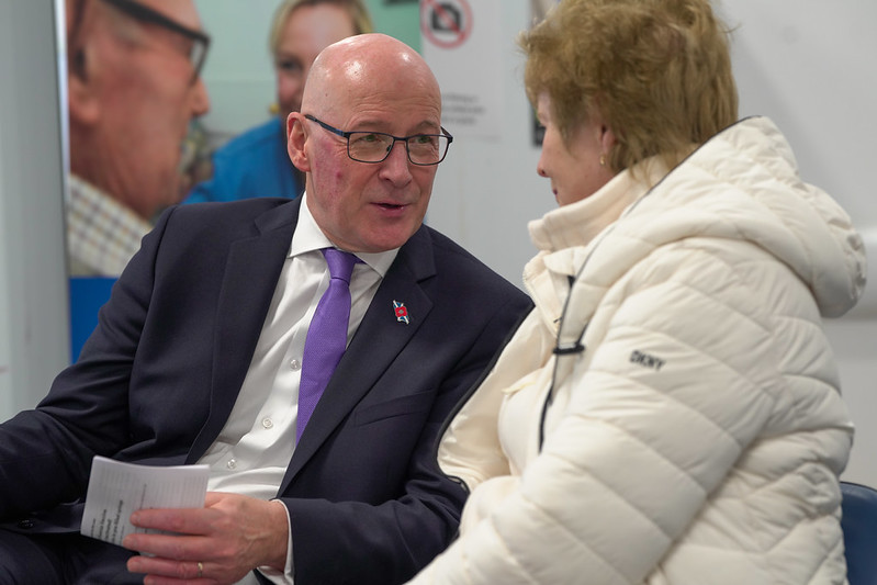 Two people engaged in a conversation, one wearing a dark suit with a purple tie and holding papers, the other in a white puffer jacket.