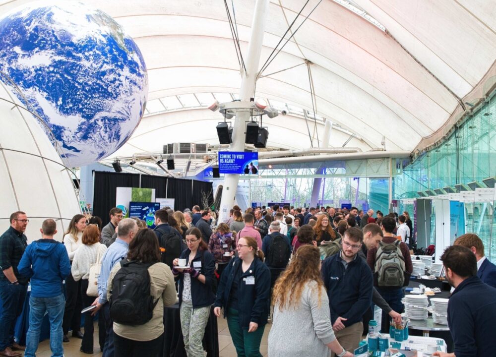 Crowded indoor event with diverse attendees networking under a large globe model suspended from the ceiling.