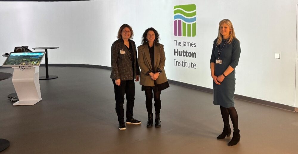 Three women standing in a modern exhibition space next to a large wall displaying The James Hutton Institute logo.