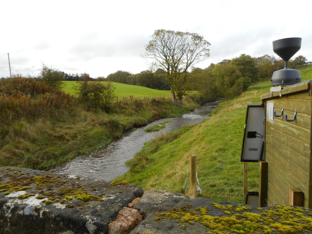 A narrow stream winds through a green countryside landscape with moss-covered rocks in the foreground and a water monitoring device on a wooden structure to the right.