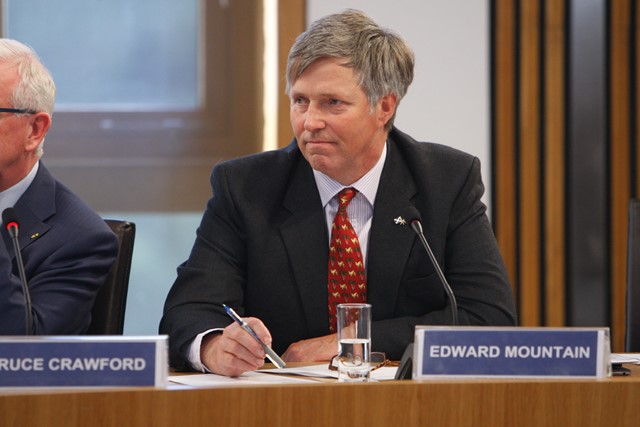 Man in a suit and red patterned tie seated at a conference table with a microphone and nameplate reading Edward Mountain.